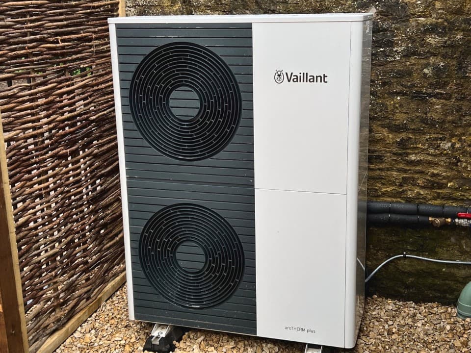 A12kW aroTHERM plus air source heat pump unit from Vaillant. The heat pump is infront of a brick wall and next to a wicker wall.