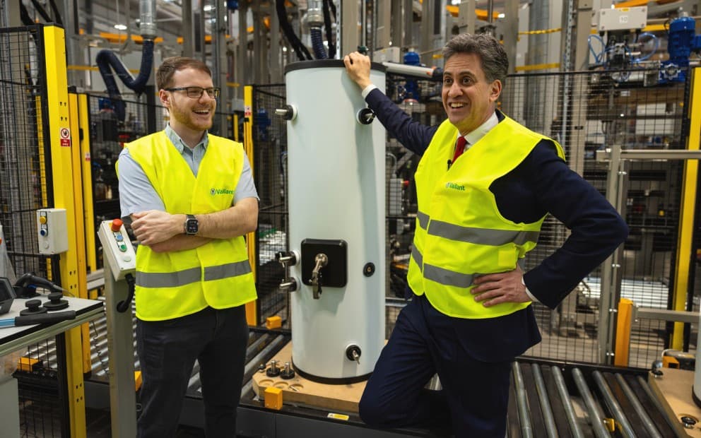 Two men in vests stand beside a large hot water cylinder, engaged in conversation.