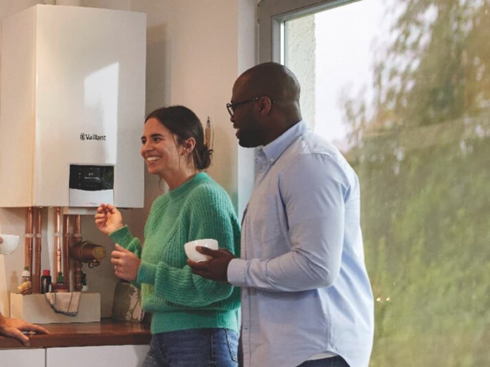 A Vaillant installer talking to two homeowners. A Vaillant gas boiler is on the wall with other kitchen utensils in the background.
