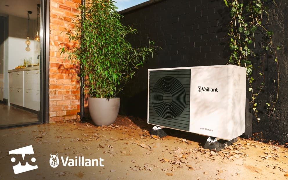 A heat pump unit outside in front of bushes and next to a brick wall.