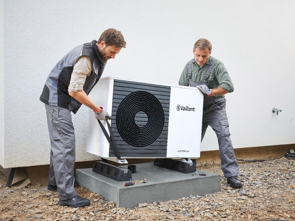 Two people lowering a heat pump unit to the ground using tools.
