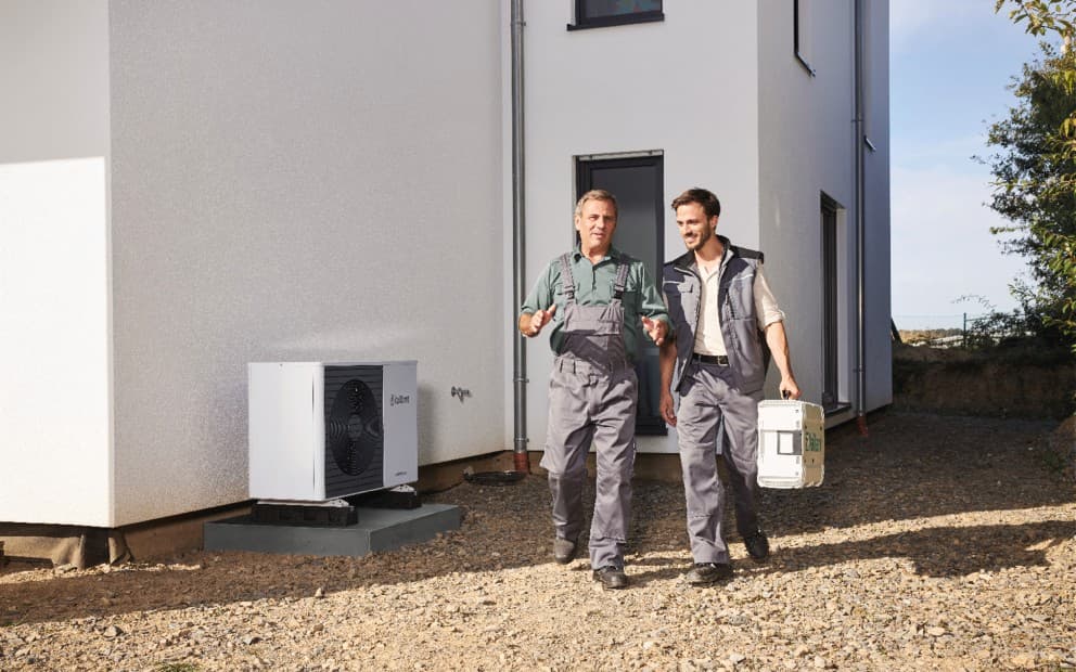 Two installer walking and talking in front of a house. There is a heat pump next to them.