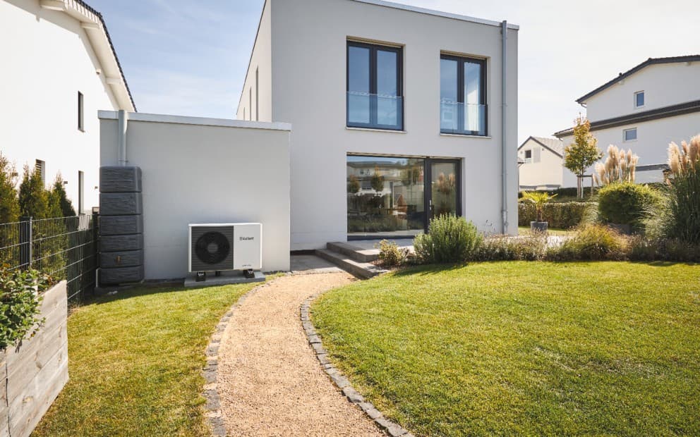 Heat pump unit outside a large white house with windows. In front of the heat pump is a garden path and grass in the garden.