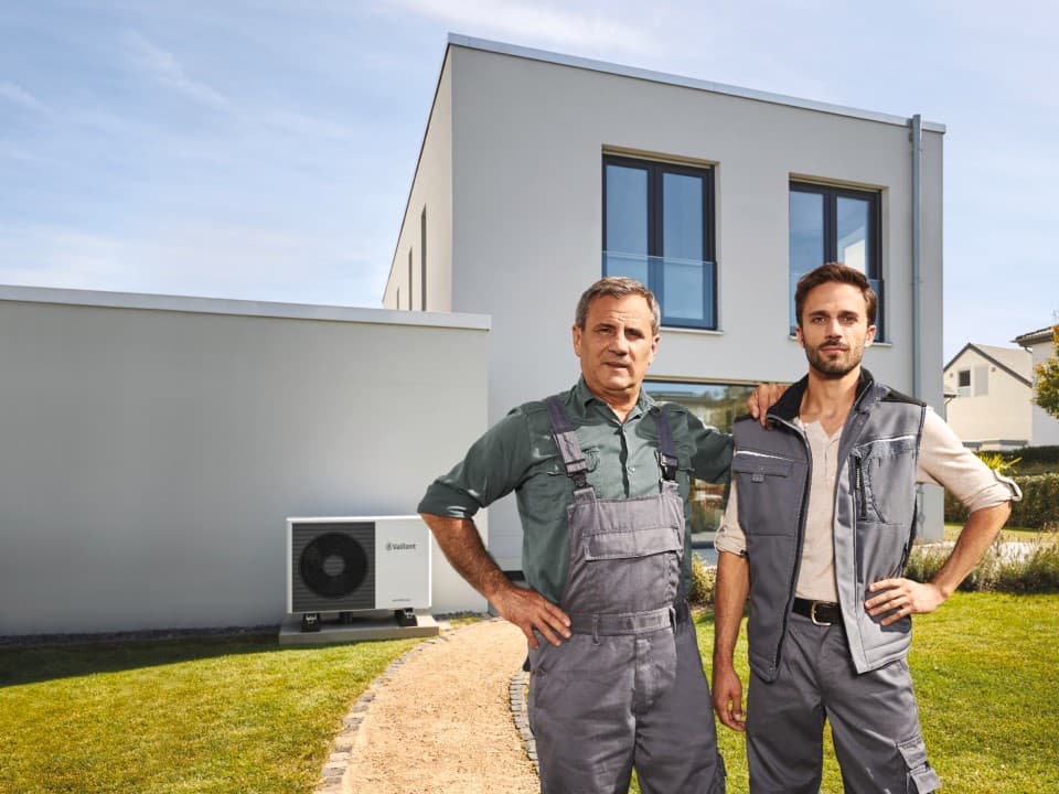 two installers standing in front of a white house and a heat pump unit