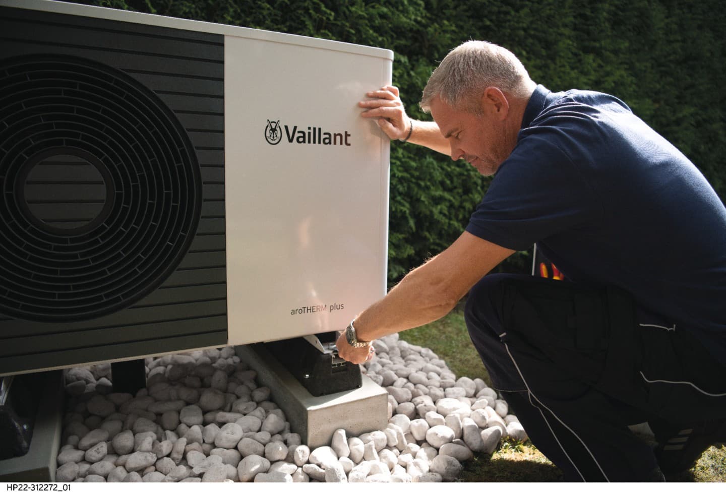 Man putting the top panel on the heat pump unit. On the ground there is a open tool box with the Vaillant logo. A bike is in the garage.