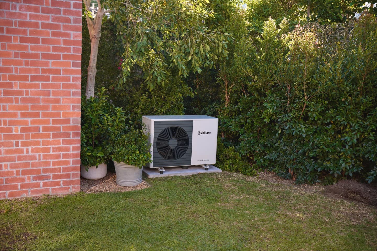 Heat pump unit behind a bush and in front of grass. There is a brick wall to the left.