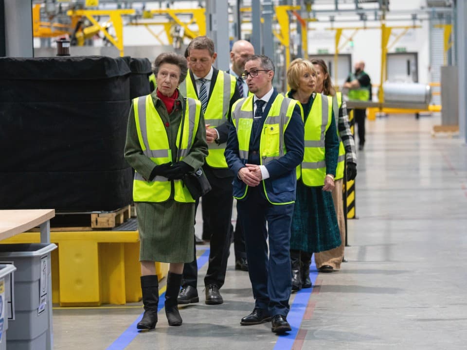HRH Princess Anne talking to Joe Dunn through Vaillant's cylinder plant wearing a hi-vis jacket. There is a line of people behind them also walking.
