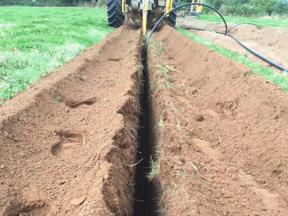 A tractor is digging a trench in a grassy field, with soil being displaced and forming a small mound beside it.