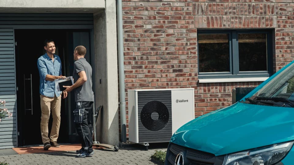 Two men in the doorway shaking hands. There is a heat pump outside the wall and in the foreground, the front of a van.