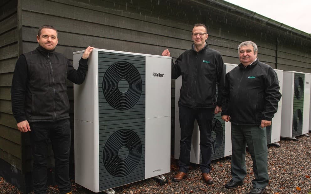 Three men standing next to a heat pump unit with other units in the background.